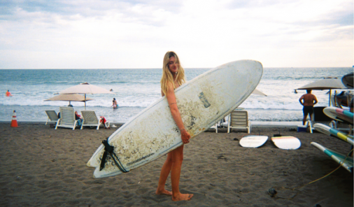 Girl Standing with Surfboard in front of the ocean with the sun shining in the background