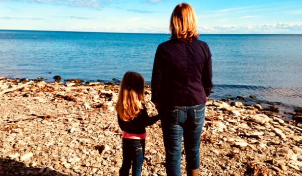 a mom holds hands with her young daughter as they look out over a blue sky and lake